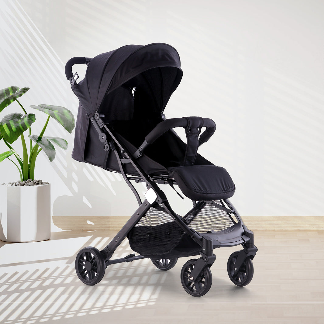 A sleek black baby stroller from the Urban Glide Collection positioned on light wooden flooring against a white wall. The stroller features an all-black design with a dark canopy, black fabric seat, and black frame with small wheels. Natural light creates geometric shadows across the scene